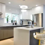 Modern kitchen with white upper cabinets, gray lower cabinets, stainless steel fridge, range hood, island with sink, and white stools with gold bases.