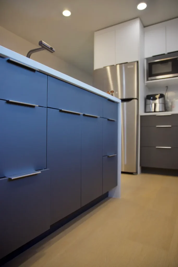 Modern kitchen interior with blue lower cabinets, stainless steel refrigerator, white upper cabinets, a built-in microwave, and light wood flooring.