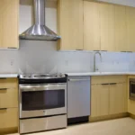 Modern kitchen with light wood cabinets, stainless steel oven, range hood, dishwasher, microwave, sink, and light wood floor.