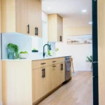 Modern kitchen with light wood cabinets, white countertop, stainless sink, plants, fruit, sliding door, wood floor, and dining area.