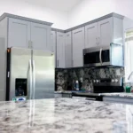 Modern kitchen with gray cabinets, stainless fridge, microwave, stove, and white marble countertops and backsplash with gray veining.