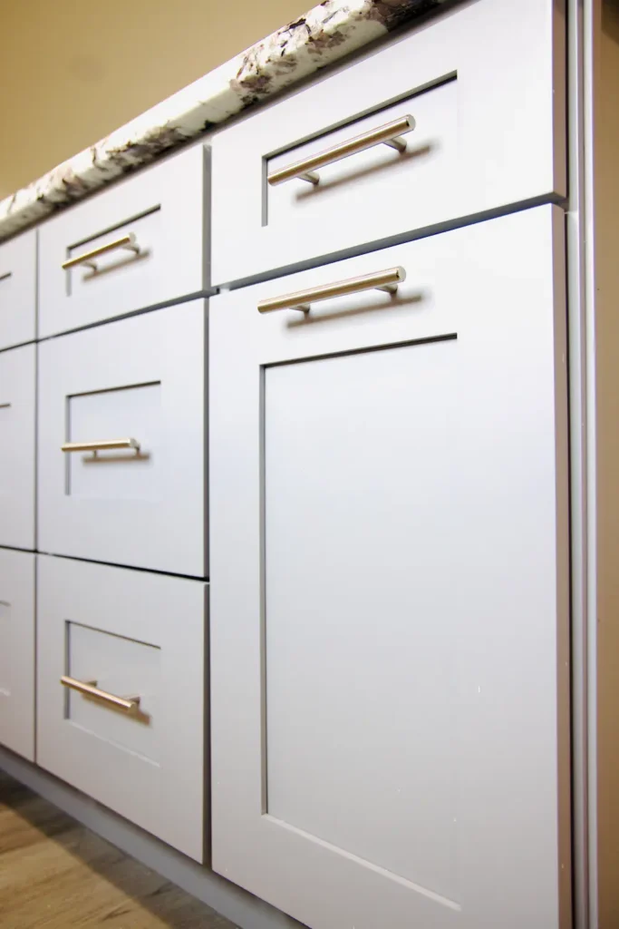 Close-up of white cabinets with metal handles under marble countertop with gray veining and wood floor.