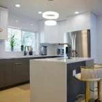 Modern kitchen with white upper cabinets, gray lower cabinets, stainless steel fridge, range hood, island with sink, and white stools with gold bases.