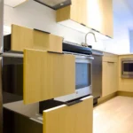 Modern kitchen with light wood cabinets, open drawers, a stainless steel oven, range hood, and light wood floor.